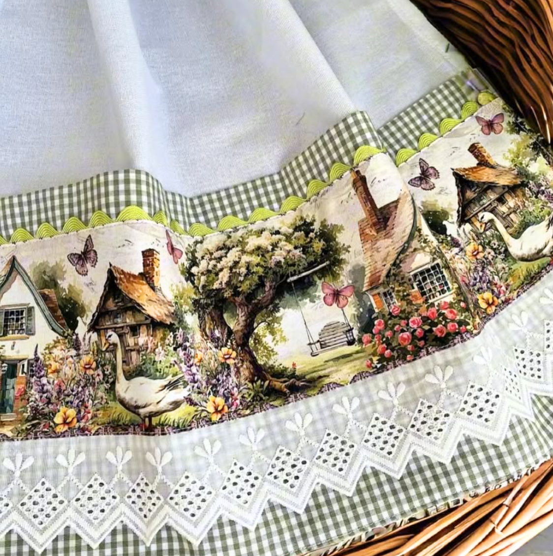 White tea towel draped over a wicker basket, showing geese, cottages, butterflies and a garden swing above white eyelet lace and sage green gingham.