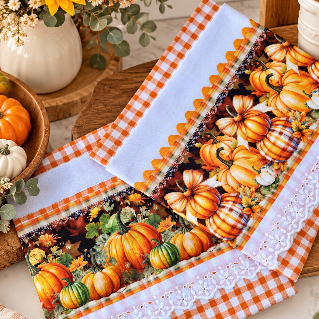 Two white tea towels with orange and green pumpkin prints on dark panels, orange gingham borders, and white eyelet lace, styled in a fall kitchen.