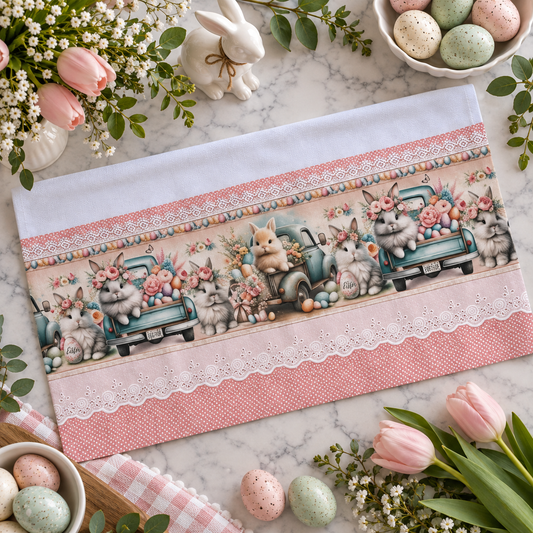 Easter Bunny Truck kitchen tea towel 20x30 flat lay on marble surface showing full print panel with fluffy bunnies riding vintage teal trucks loaded with pastel Easter eggs and pink roses, pink polka dot trim with white eyelet lace border, surrounded by pink tulips, speckled pastel eggs, white ceramic bunny figurine, and spring eucalyptus greenery