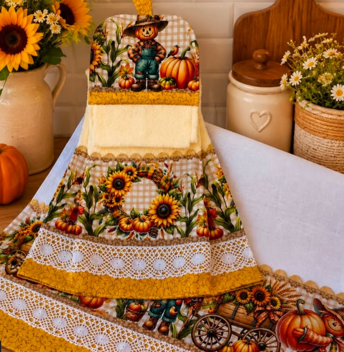 Fall kitchen towels with scarecrow, pumpkin, and sunflower prints, white lace and mustard yellow trim, displayed on a kitchen counter.