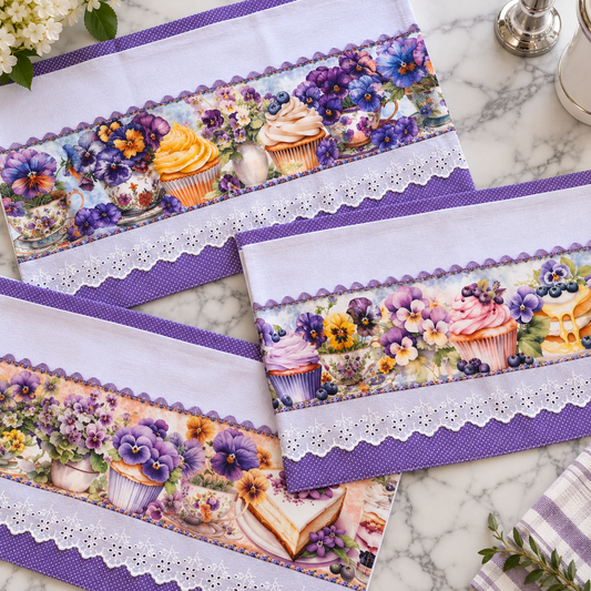 Pansy Garden kitchen tea towel set 3-piece flat lay on marble surface showing three coordinating purple pansy and cupcake print towels with purple polka dot trim and white eyelet lace border, each with slightly different pansy and dessert scene arrangement